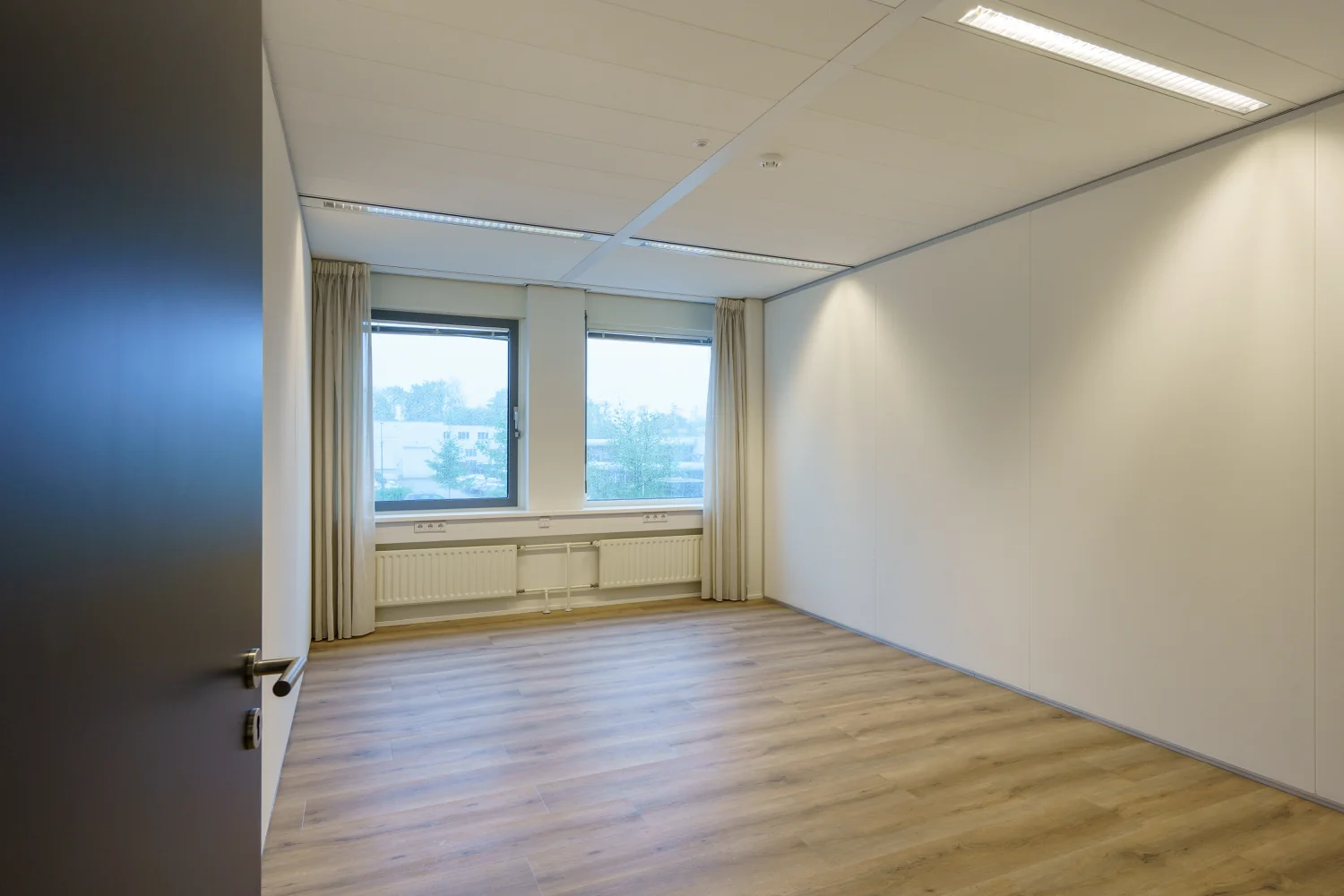 Lege, lichte kantoorruimte aan de Baarnsche Dijk met houten vloer en twee ramen met uitzicht op een bedrijventerrein.